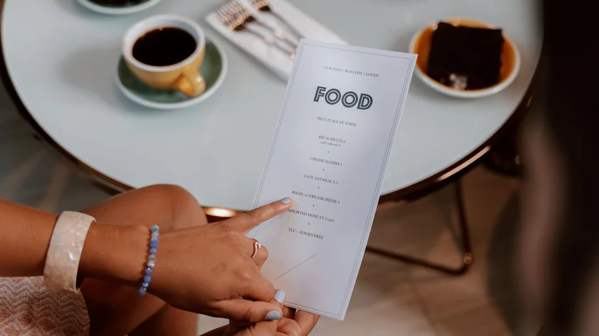 Des mains consultent une carte papier blanche devant une table avec un café et un dessert.