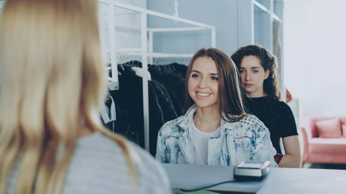 Deux clientes échangent avec une vendeuse au comptoir d’une boutique de vêtements.