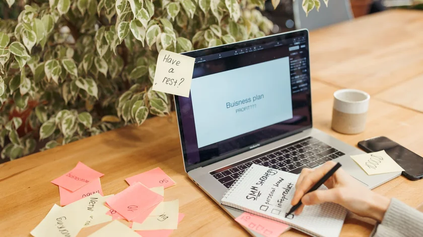 Une main prend des notes près d’un ordinateur portable et de papiers adhésifs roses.