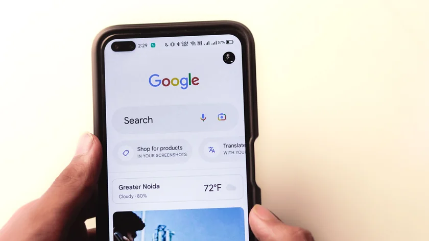 Des mains tiennent un smartphone affichant l’écran d’accueil Google sur un fond clair.