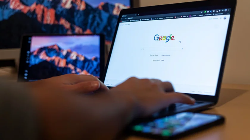Des mains sur le clavier d’un ordinateur portable affichant la page de recherche Google, avec un téléphone posé devant.